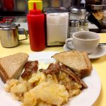 2 Egg platter w Sausage. This picture depicts an order of two eggs Fried with on fried potatoes, sausage and Toast cut in half on top of a platter with a cuppa coffee on the side, served on Johnny’s counter tap