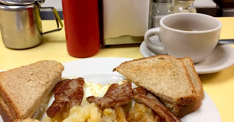 2 Egg platter w Sausage. This picture depicts an order of two eggs Fried with on fried potatoes, sausage and Toast cut in half on top of a platter with a cuppa coffee on the side, served on Johnny’s counter tap
