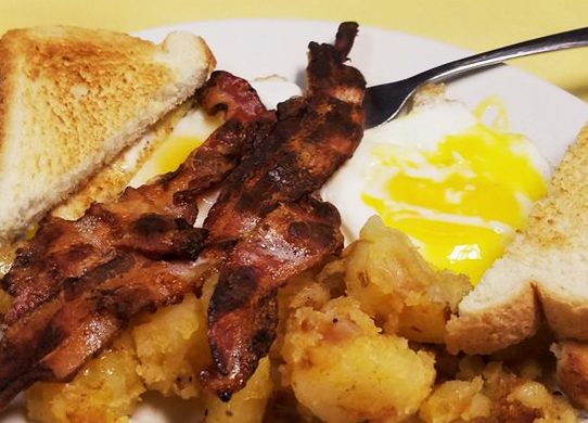 2 egg platter with bacon. This picture depicts two sunny side up eggs Served with home fried potatoes on the side, topped with three pieces of bacon and two slices of toast cut in half placed on top.