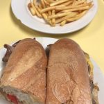 Cheese Steak Hero. This picture depicts a sandwich that consists of sliced steak, peppers and onions, and melted American cheese on a sub that is cut in half served on a white platter accompanied by a side of french fries for an extra charge on top of Johnys counter