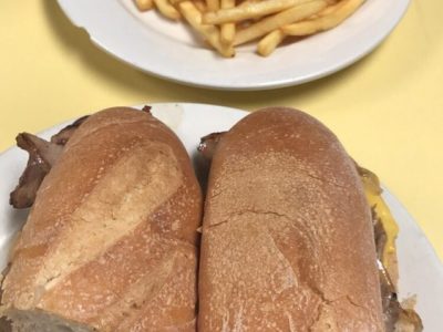 Cheese Steak Hero. This picture depicts a sandwich that consists of sliced steak, peppers and onions, and melted American cheese on a sub that is cut in half served on a white platter accompanied by a side of french fries for an extra charge on top of Johnys counter