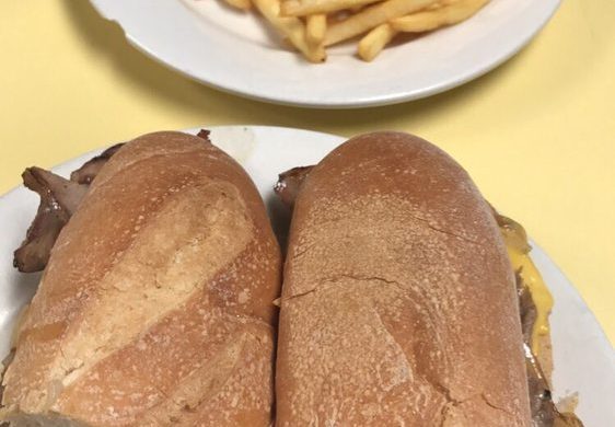 Cheese Steak Hero. This picture depicts a sandwich that consists of sliced steak, peppers and onions, and melted American cheese on a sub that is cut in half served on a white platter accompanied by a side of french fries for an extra charge on top of Johnys counter