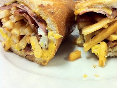 Curious George. This picture shows a sandwich that consists of three Fried eggs, Bacon and sliced ham, Melted American cheese and french fries on a semolina hero that is cut in half and served on a White platter on top of Johnys countertop