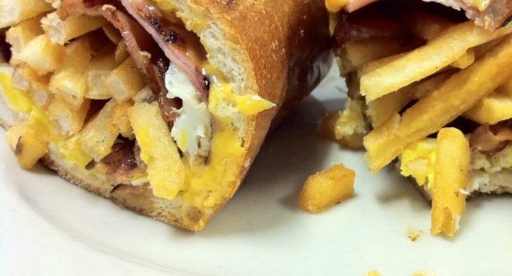 Curious George. This picture shows a sandwich that consists of three Fried eggs, Bacon and sliced ham, Melted American cheese and french fries on a semolina hero that is cut in half and served on a White platter on top of Johnys countertop