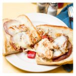 The Greco. This picture depicts an order of grilled chicken with roasted red peppers, grilled salami and melted mozzarella cheese that is cut in half and served open face on a white platter serve lunch and he’s going