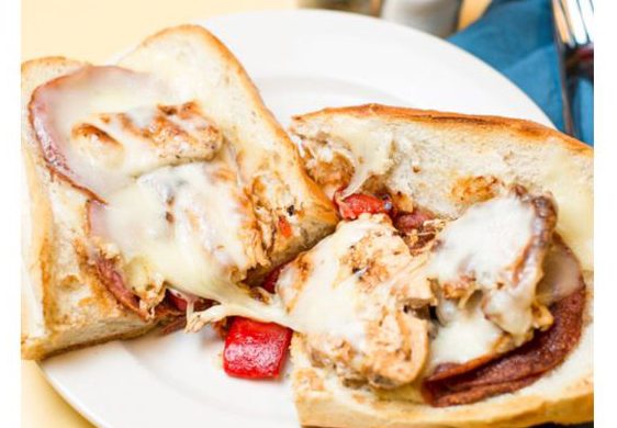 The Greco. This picture depicts an order of grilled chicken with roasted red peppers, grilled salami and melted mozzarella cheese that is cut in half and served open face on a white platter serve lunch and he’s going