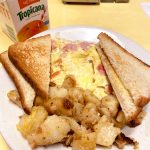 Western Omelette. This picture depicts an omelette which consists of chopped ham, peppers and onions served with home fries potatoes on the side and two slices of toast on top, served on a white planter on Johnny’s counter top