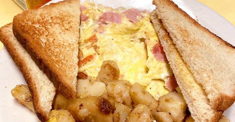 Western Omelette. This picture depicts an omelette which consists of chopped ham, peppers and onions served with home fries potatoes on the side and two slices of toast on top, served on a white planter on Johnny’s counter top