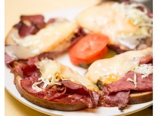 Pastrami Reuben. This picture depicts an order of grilled pastrami, homemade coleslaw Russian dressing and melted Swiss cheese on top of grills Ryebread that is cut into quarters Served open face on a white plaid or on top of Johnys countertop