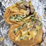 Spinach Egg Sausage and Cheese Wrap. This picture depicts a rap that consists of Two eggs, spinach, sausage and melted cheese that is cut in half ends Served open face on a piece of take out aluminum foil