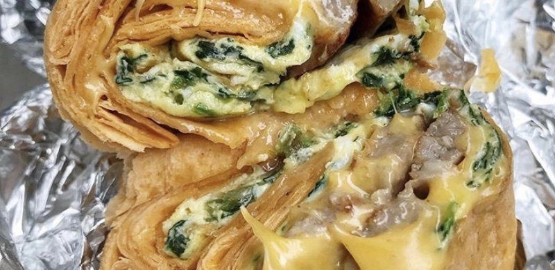 Spinach Egg Sausage and Cheese Wrap. This picture depicts a rap that consists of Two eggs, spinach, sausage and melted cheese that is cut in half ends Served open face on a piece of take out aluminum foil